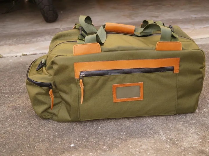 Olive canvas duffel bag with tan leather trim and a clear ID window, sitting on a concrete floor